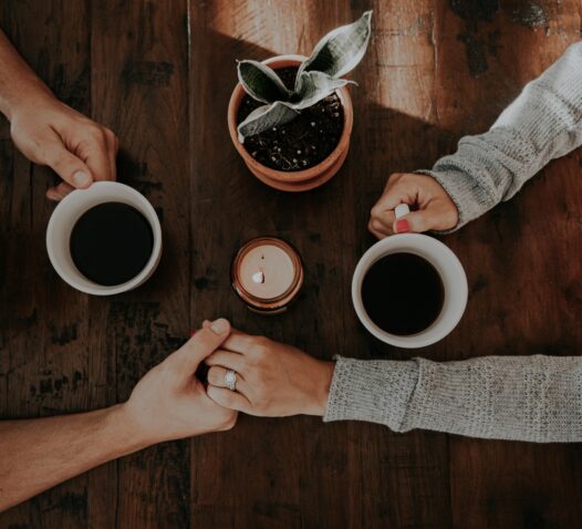 Tre händer runt en bordsskiva med två kaffekoppar och en krukväxt.